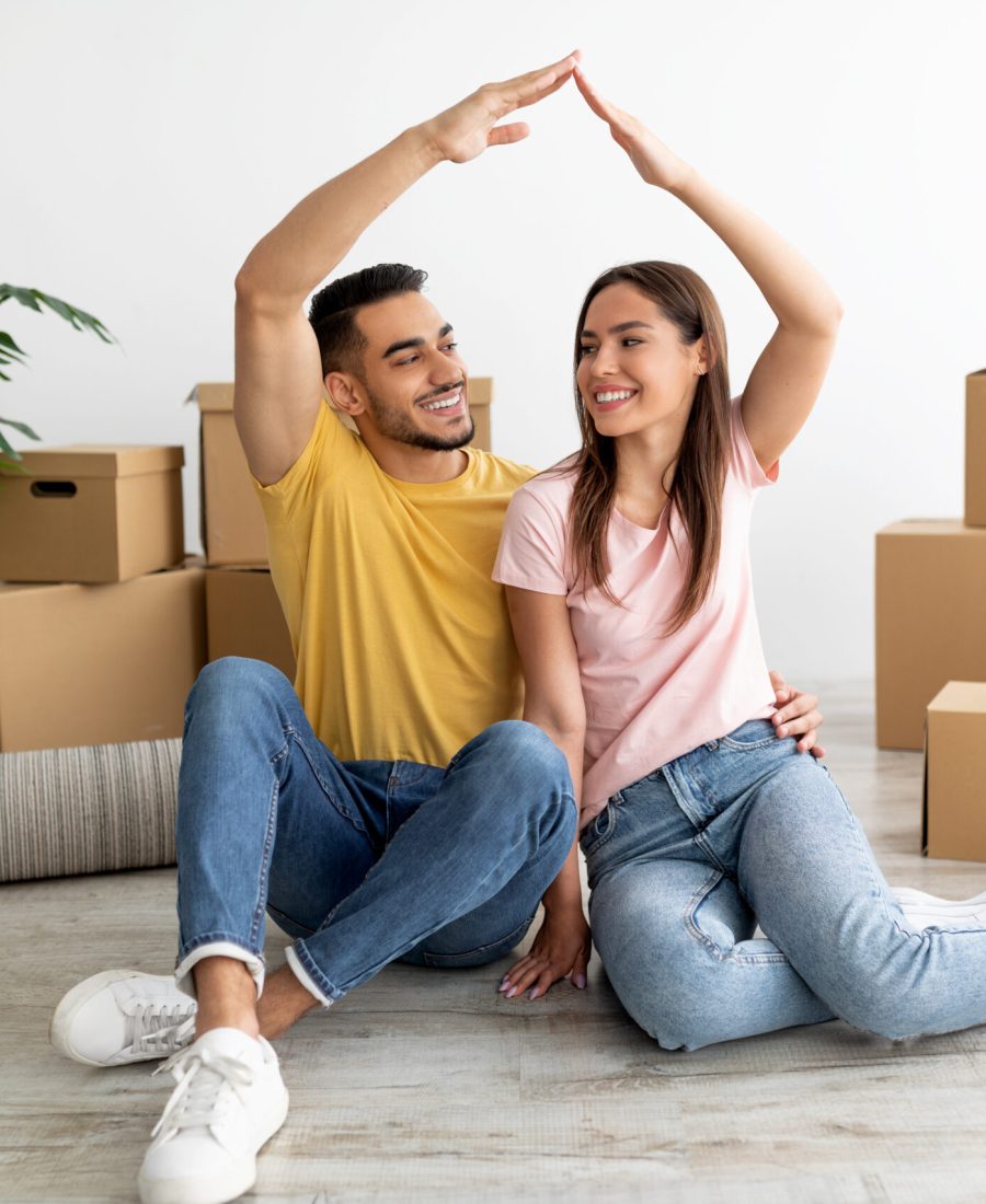 Full length of happy millennial international couple making house gesture, sitting on floor among carton boxes, moving to new apartment together. Relocation day, renting property concept